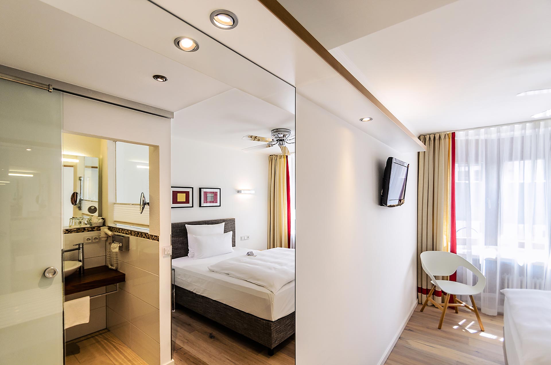 Das Hotel Restaurant Schwanen in Metzingen bietet seinen Gästen eine moderne und stilvolle Unterkunft. Das Bild zeigt ein helles und einladendes Zimmer, das mit einem komfortablen Bett, einem Flachbildfernseher und einer angenehmen Sitzgelegenheit ausgestattet ist. Die große Fensterfront sorgt für viel Tageslicht und schafft eine freundliche Atmosphäre.